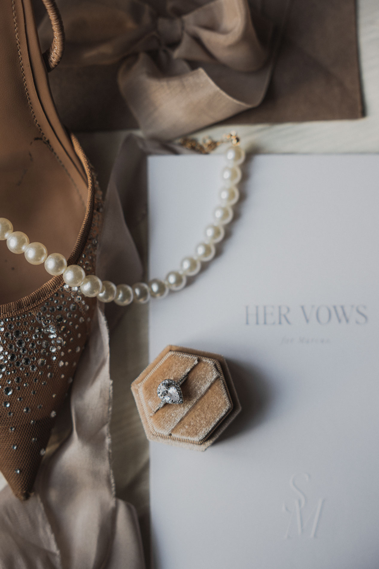 Close-up of wedding details: a velvet ring box with a teardrop-shaped engagement ring, pearl necklace, and vow booklet.