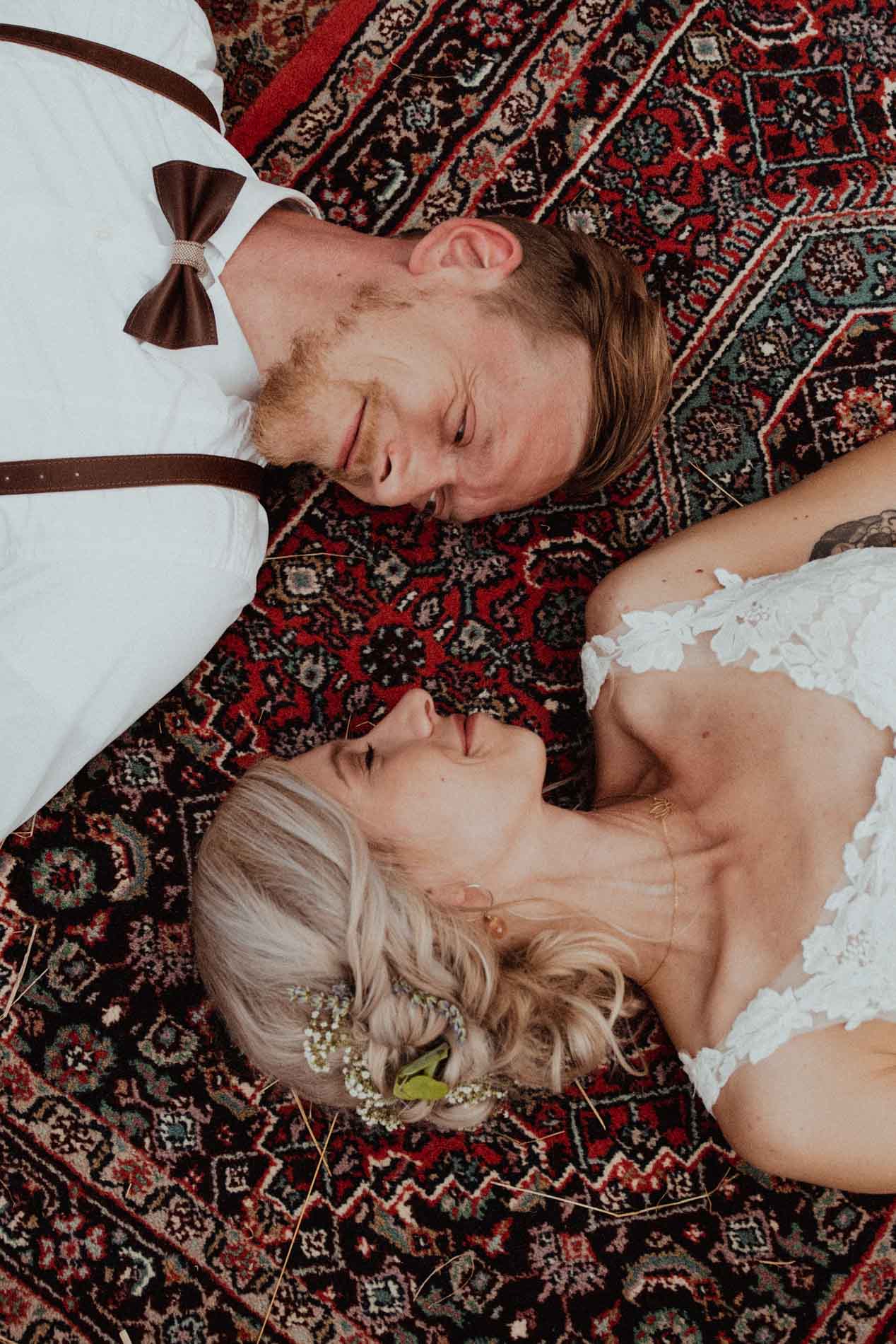 Bride and groom lying on a patterned rug, gazing into each other’s eyes and smiling after their wedding ceremony.
